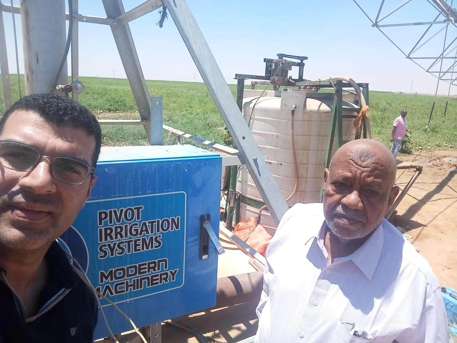 Two men beside the blue AksuPivot control panel at a pivot point in Egypt, with a water tank and green crop behind